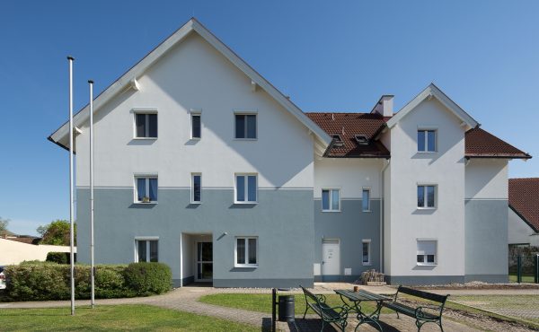 Modernes Wohnhaus mit Garten, blauer Himmel, Sitzbänke und Autos, sonniger Tag.