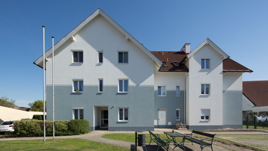 Modernes Wohnhaus mit Garten, blauer Himmel, Sitzbänke und Autos, sonniger Tag.