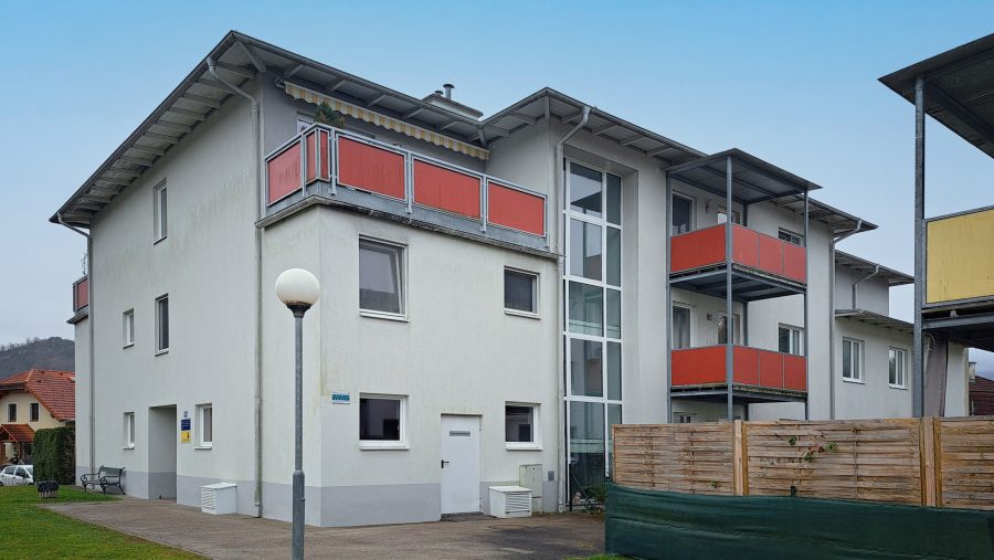 Mehrfamilienhaus mit roten Balkonen und gepflegtem Außenbereich an einem klaren Tag.