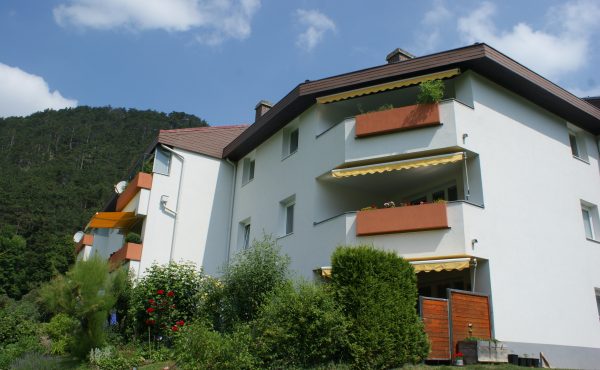 Wohnanlage mit Balkonen und Garten vor bewaldetem Hügel im Hintergrund unter blauem Himmel.