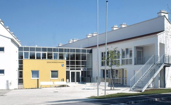 Modernes Seniorenwohnheim mit gelben und weißen Fassaden, großer Eingangsbereich und Außentreppen bei klarem Himmel.