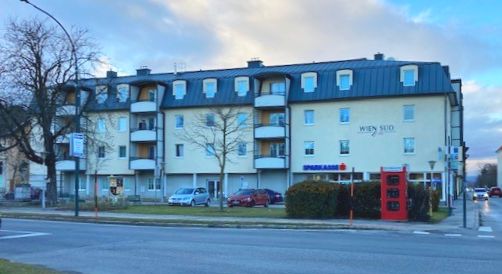 Mehrstöckiges Wohnhaus mit Balkonen und Telefonzelle vor einer Straße bei bewölktem Himmel.