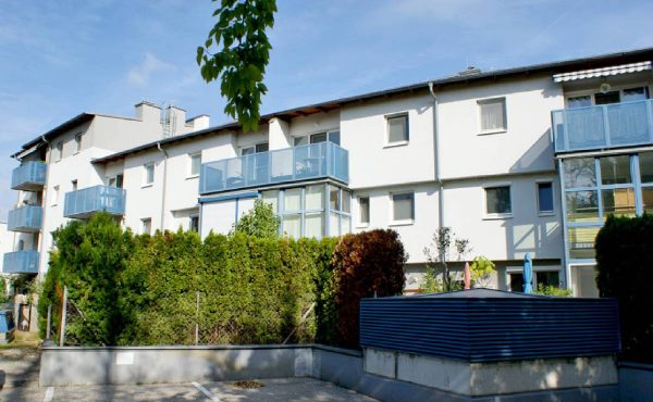 Mehrfamilienhaus mit Balkonen und Gärten, sonniger Tag, klare blauer Himmel.