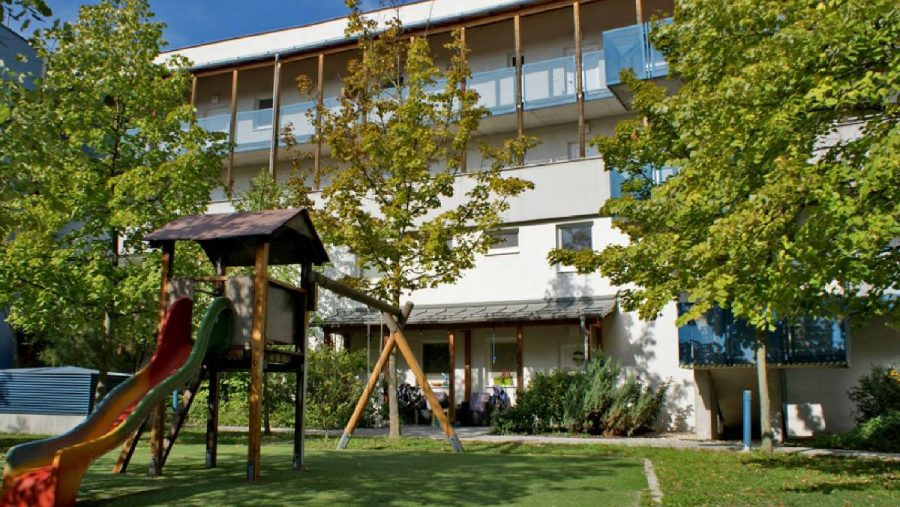 Wohngebäude mit Spielplatz im Vordergrund, umgeben von grünen Bäumen an einem sonnigen Tag.