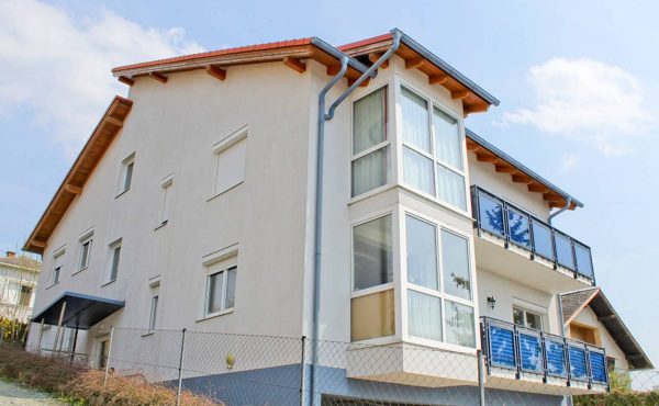 Modernes Wohnhaus mit großem Balkon und Fenstern, umgeben von einem Zaun, bei Sonnenschein.