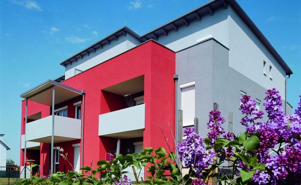 Modernes Wohngebäude mit roter und grauer Fassade, umgeben von lila blühenden Pflanzen unter blauem Himmel.
