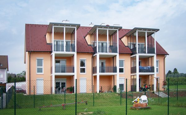 Wohngebäude mit Balkonen und Garten vor bewölktem Himmel.
